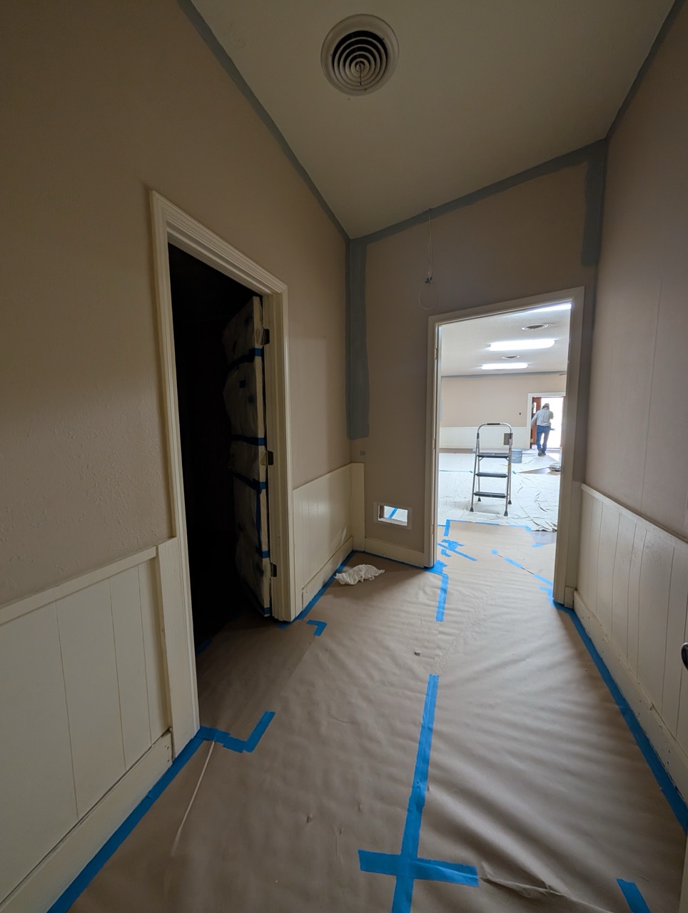 Hallway under renovation with floor covered, taped edges, and a ladder in the adjoining room.