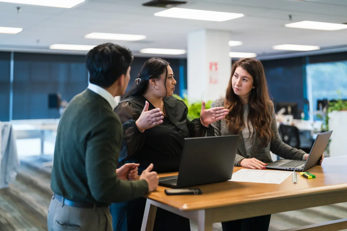 Three colleagues talking over laptops at an office table