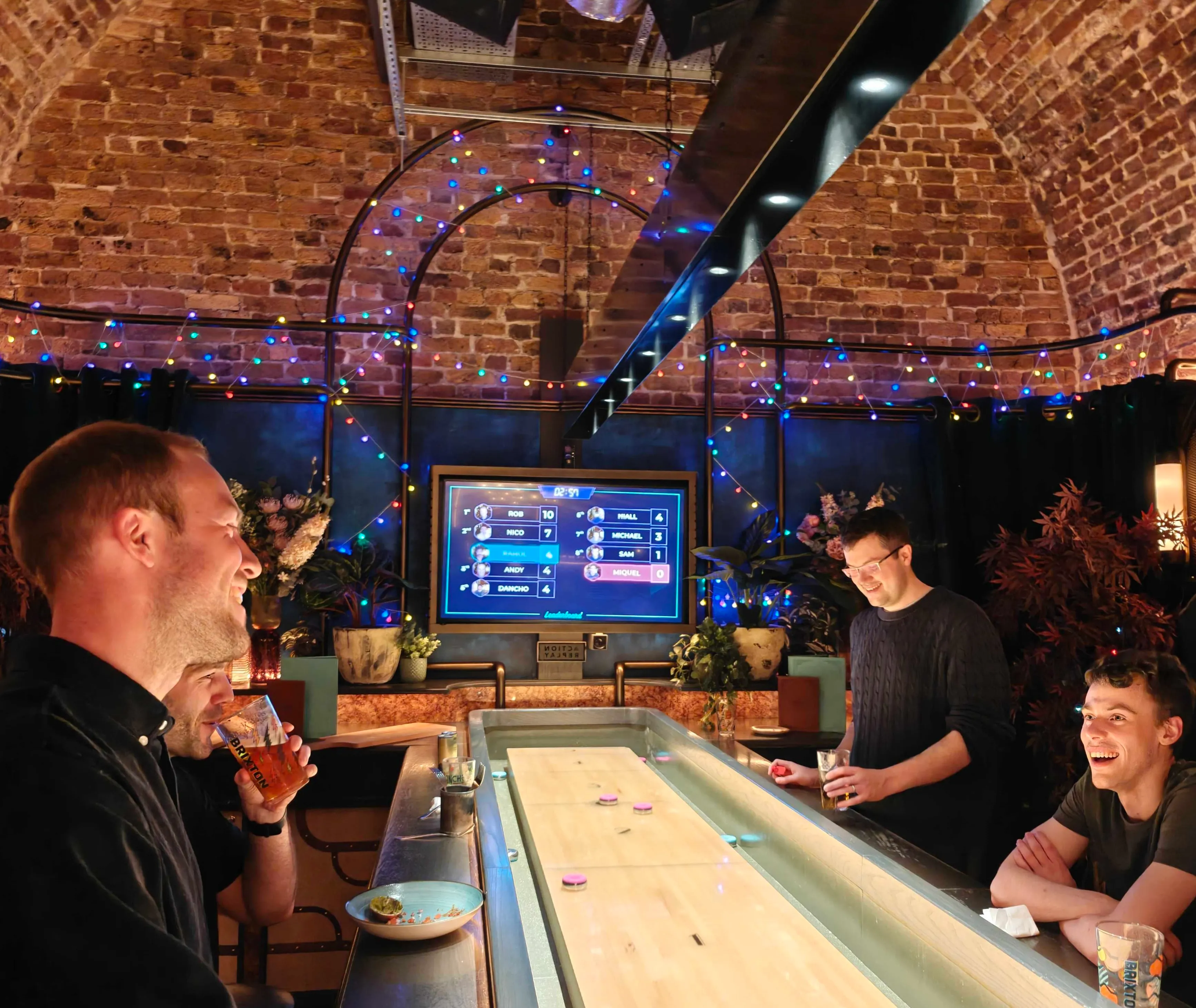 Four men enjoying drinks and playing shuffleboard in a warmly lit bar with exposed brick walls and colorful string lights.