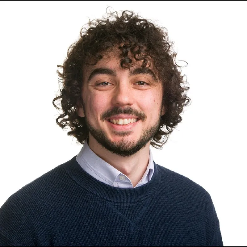 Smiling man with curly brown hair, beard, wearing a navy blue sweater over a light blue collared shirt in front of a white background.