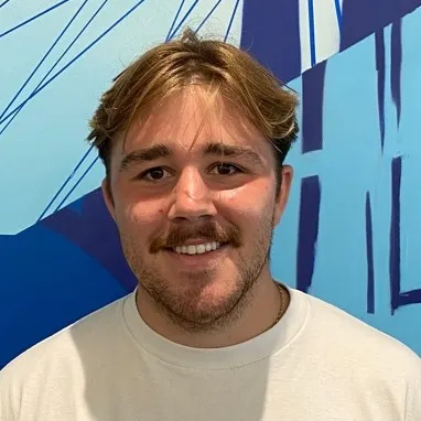Smiling young man with short light brown hair and a mustache wearing a white shirt in front of a blue abstract wall.