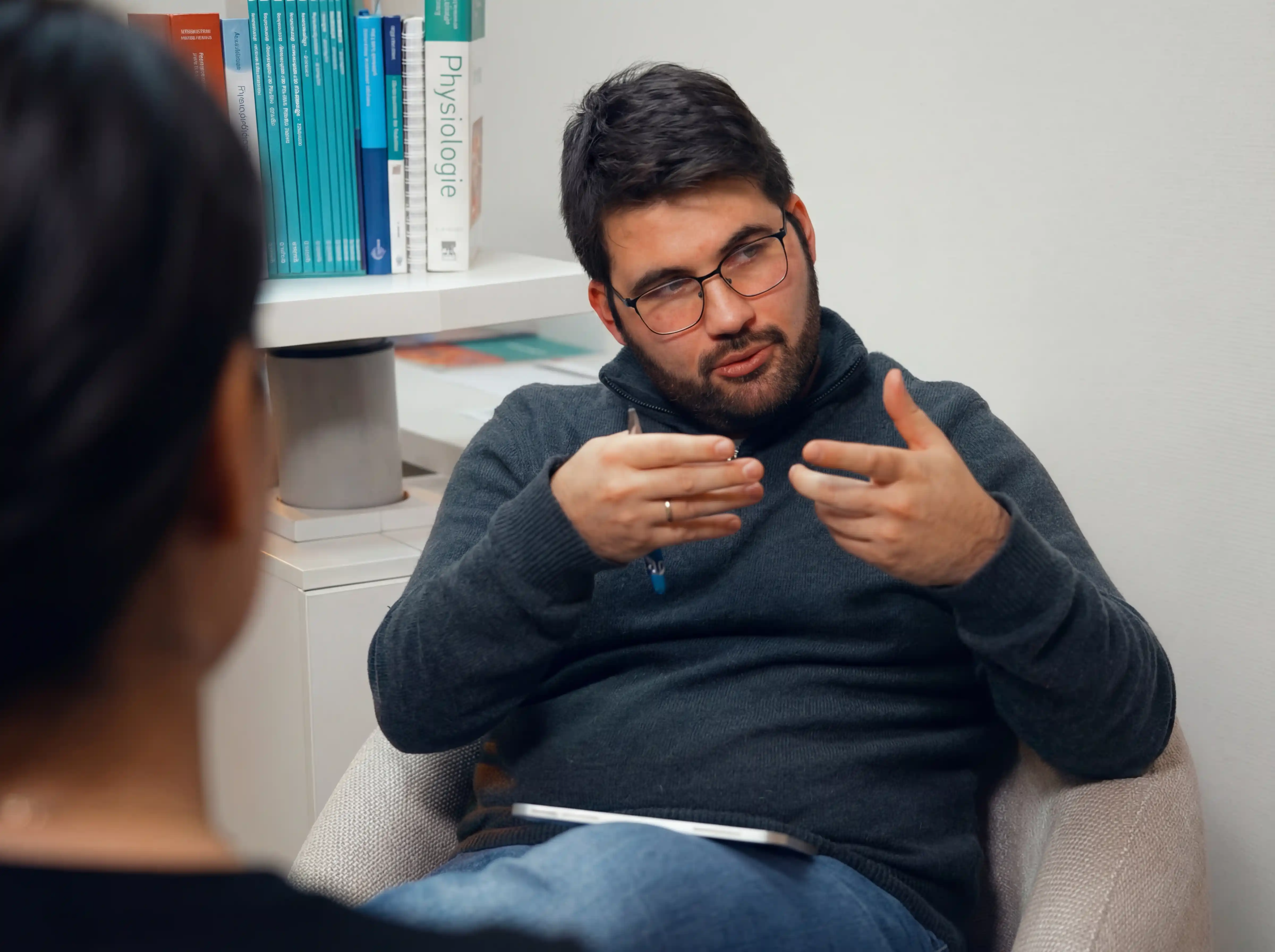 Mann mit Brille, der in einem Stuhl sitzt und mit einer anderen Person in einem Gespräch gestikuliert.