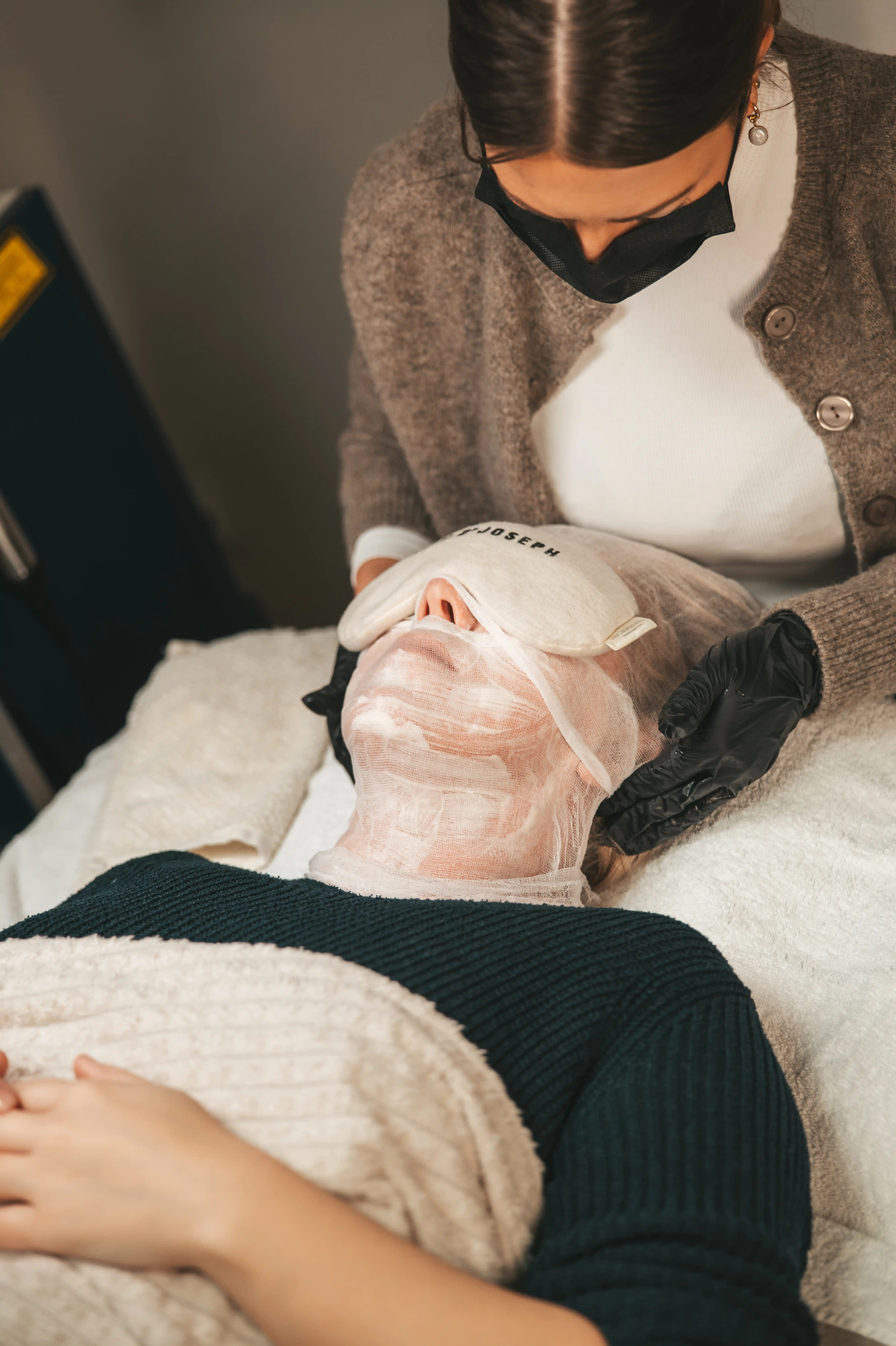 Kosmetikerin mit schwarzer Maske und Handschuhen behandelt eine liegende Kundin mit einer Gesichtsmaske aus Mullverband und einer Augenbinde.