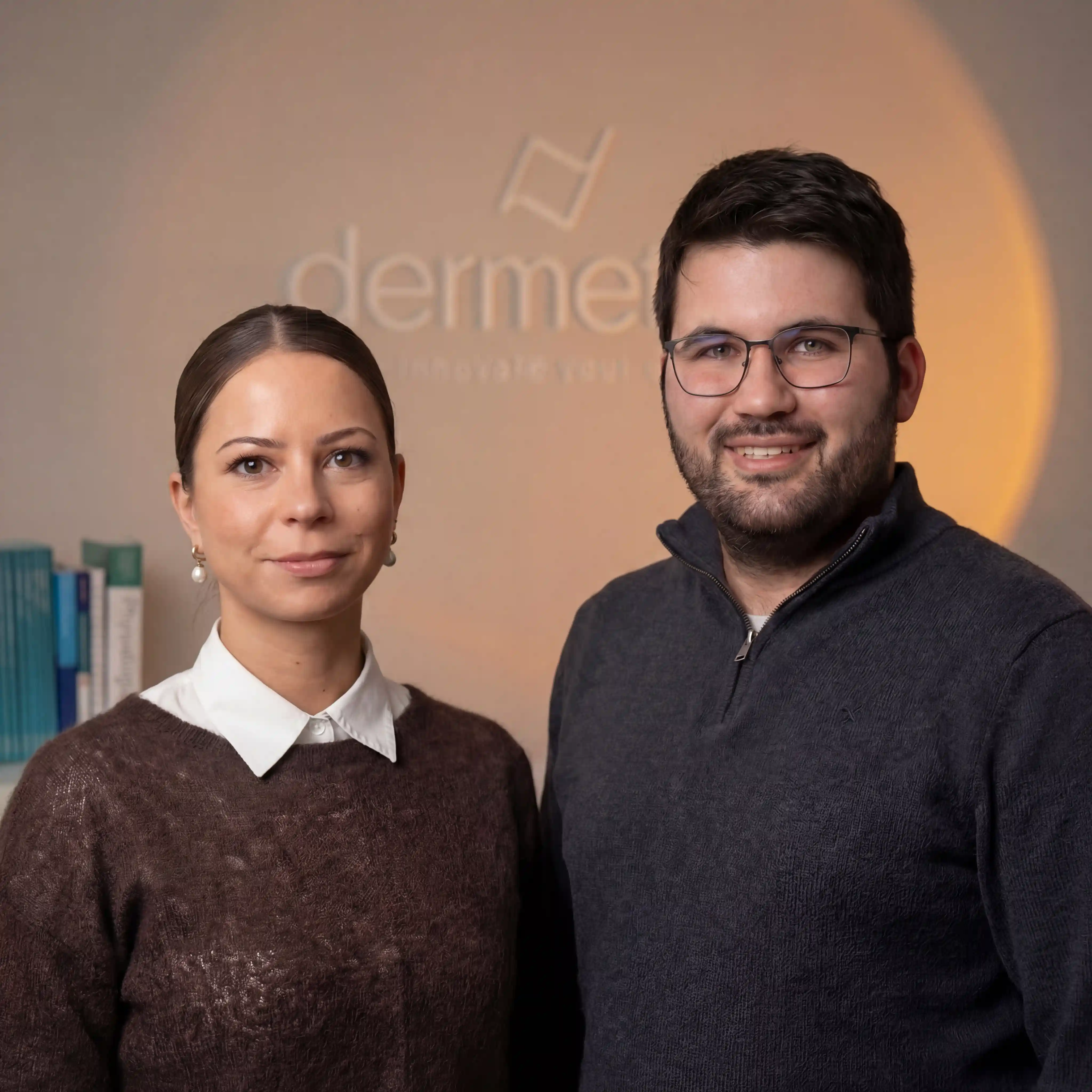 Porträt eines lächelnden Mannes mit Brille neben einer Frau mit braunem Pullover und weißem Hemdkragen vor einer Wand mit dem Schriftzug 'dermetics'.