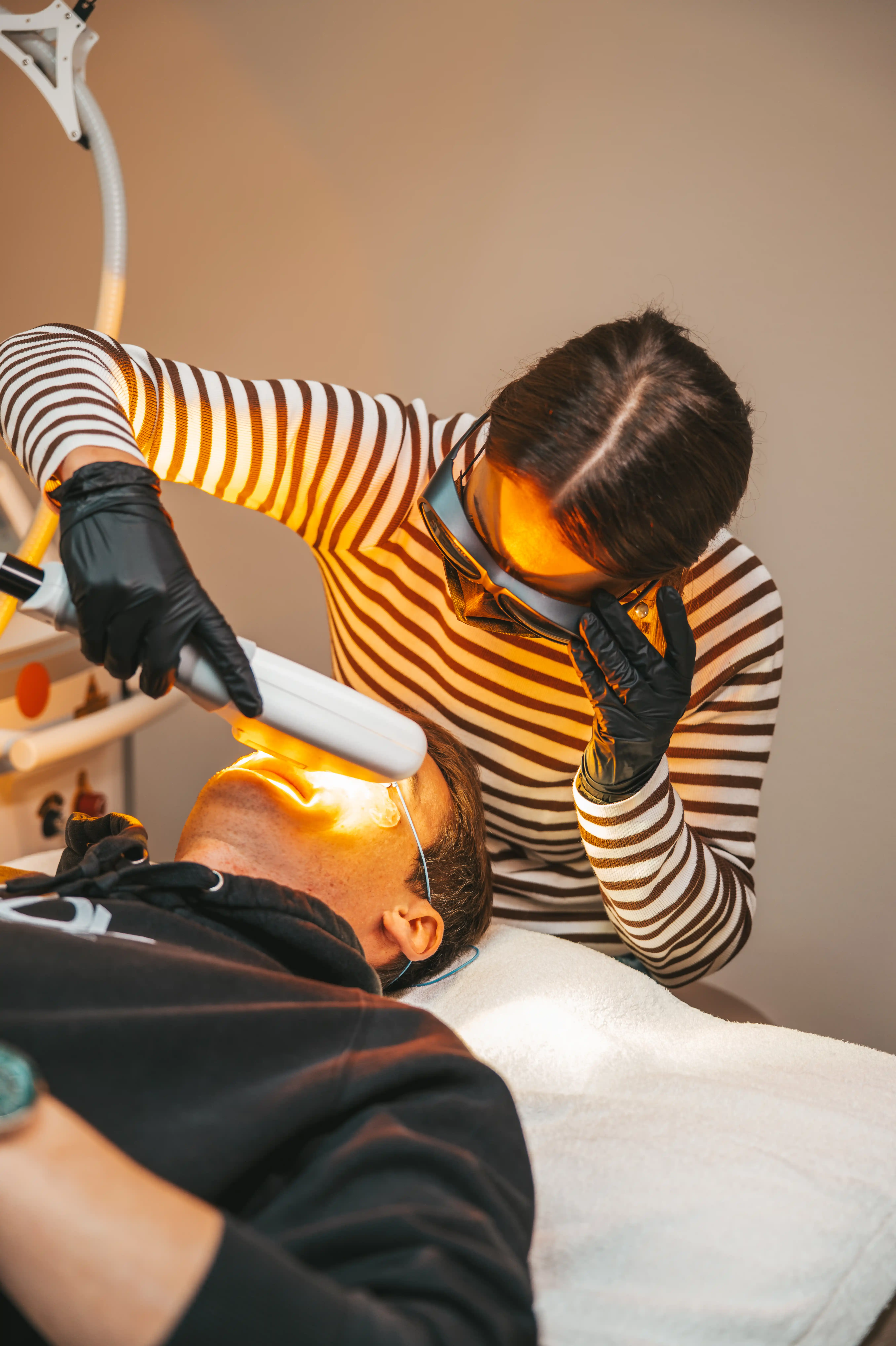 Kosmetikerin mit schwarzer Schutzbrille und Handschuhen führt eine Laserbehandlung im Gesicht eines liegenden Mannes durch.