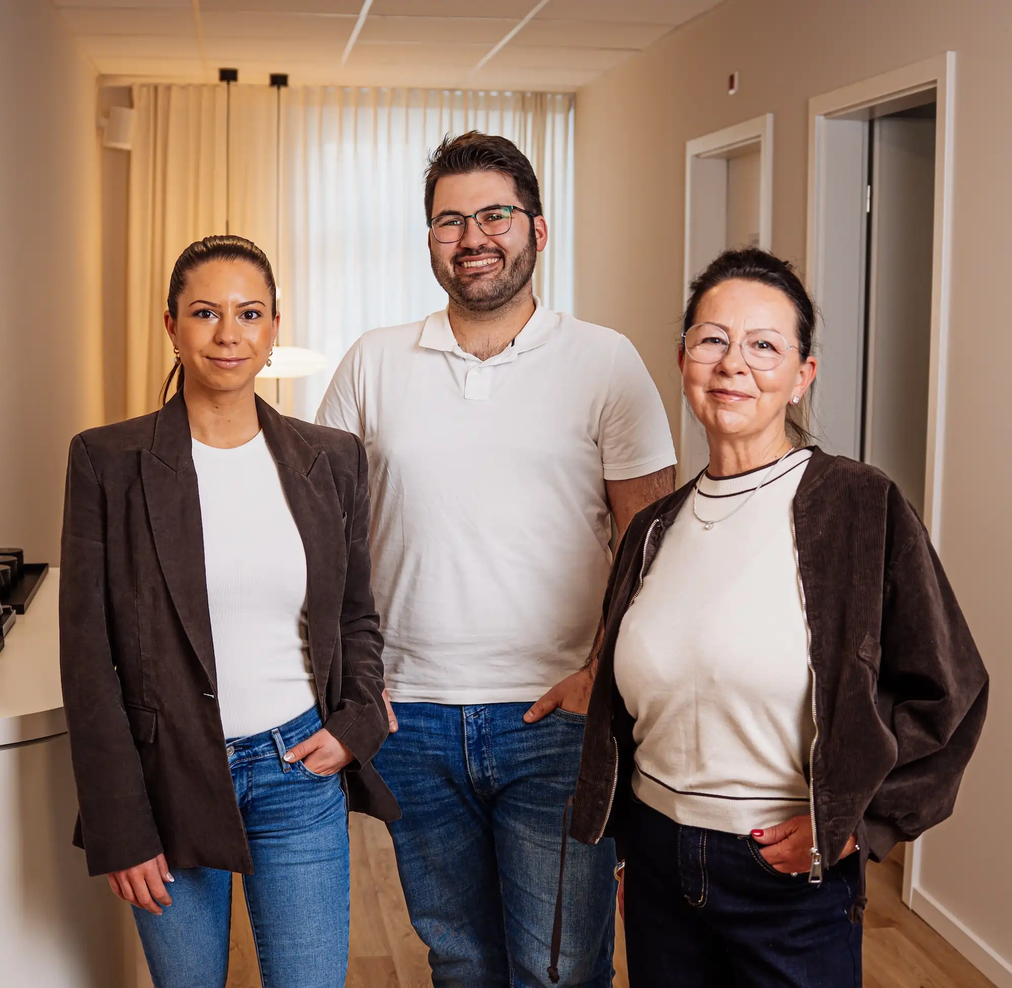 Drei lächelnde Personen im Flur, zwei Frauen tragen dunkle Jacken und Jeans, der Mann in der Mitte ein weißes Poloshirt.
