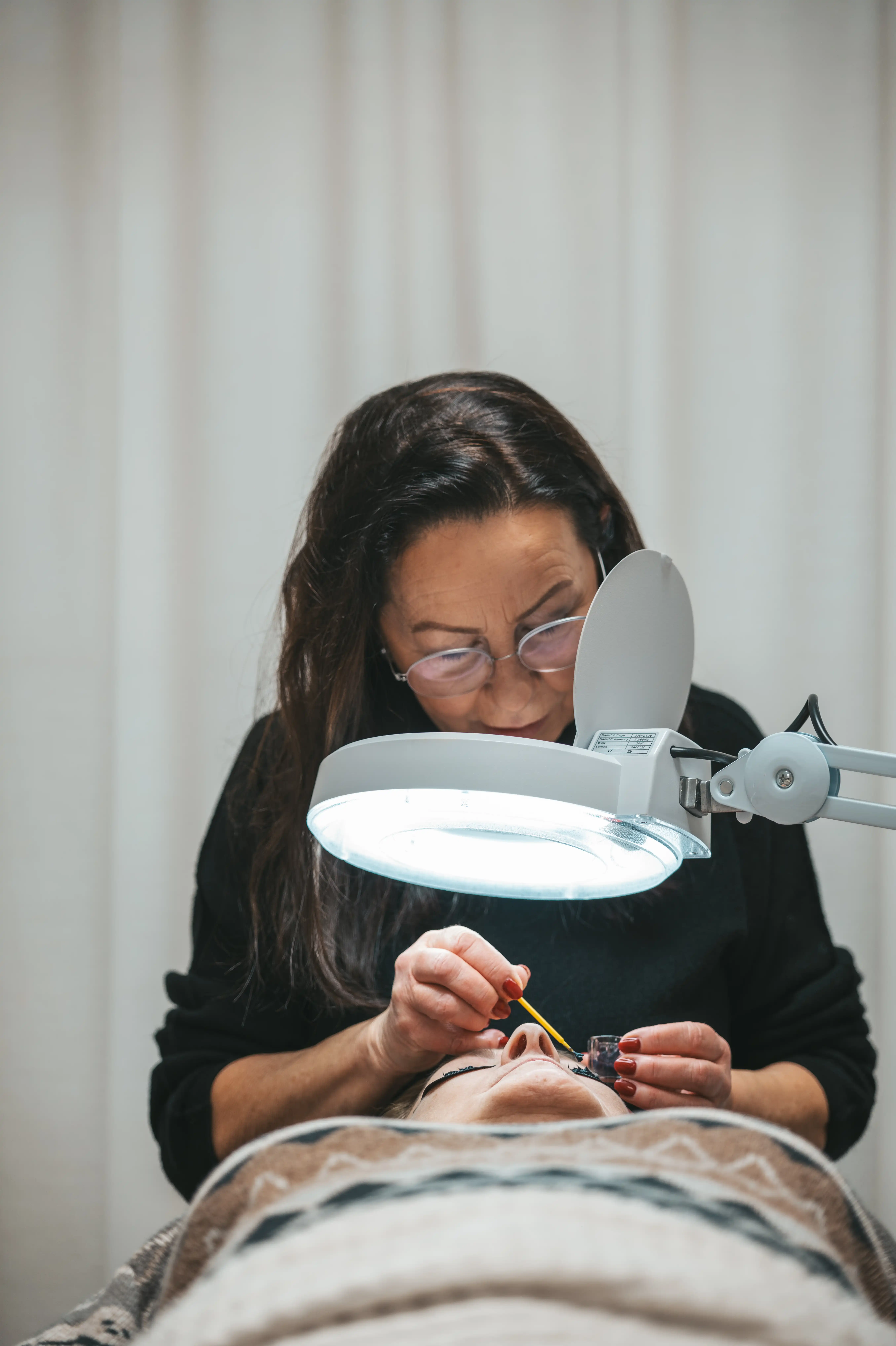 Kosmetikerin trägt Wimpernextensions bei einer liegenden Kundin unter einer Beleuchtung auf.