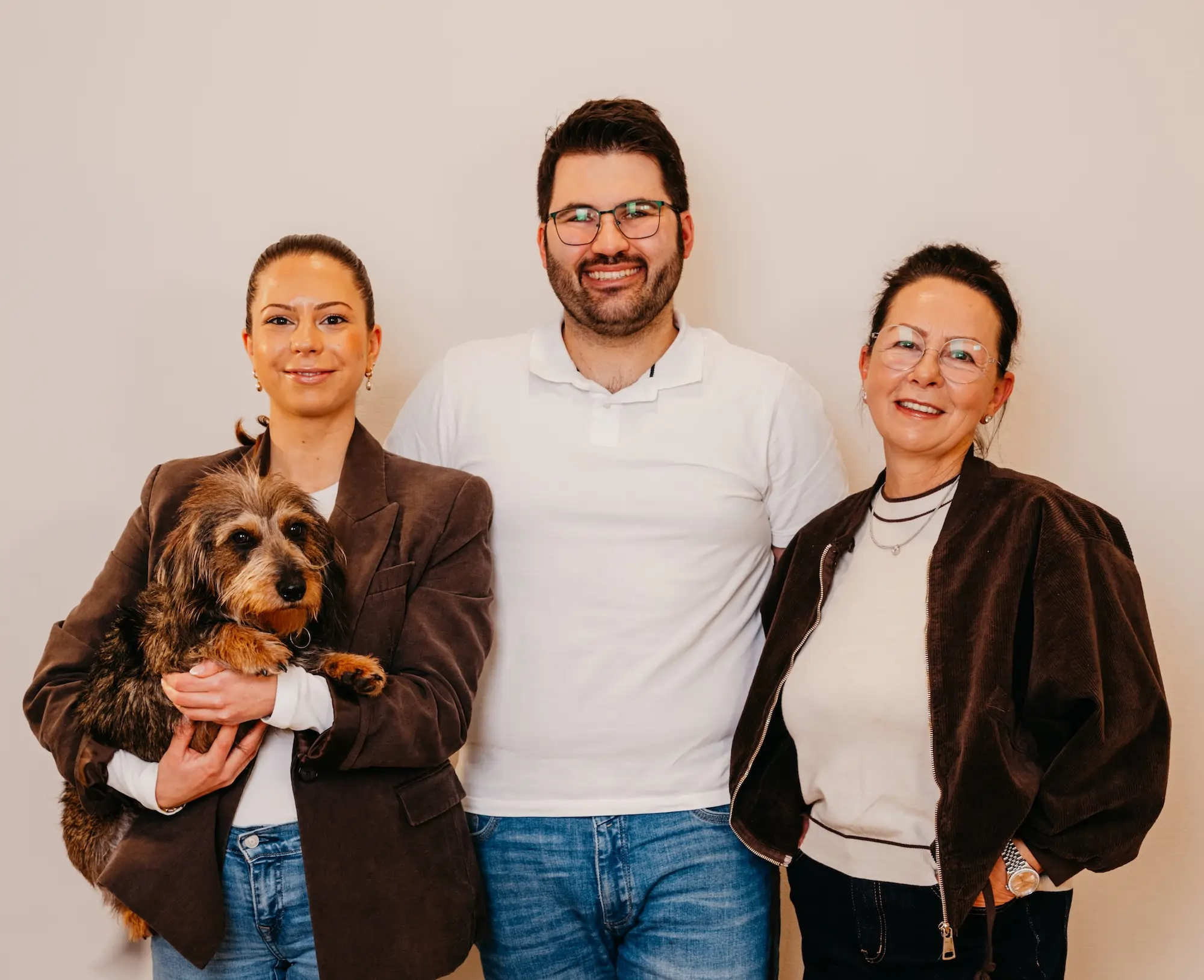 Drei lächelnde Personen, zwei Frauen und ein Mann, vor einem hellen Hintergrund, eine Frau hält einen kleinen, braun-schwarzen Hund.