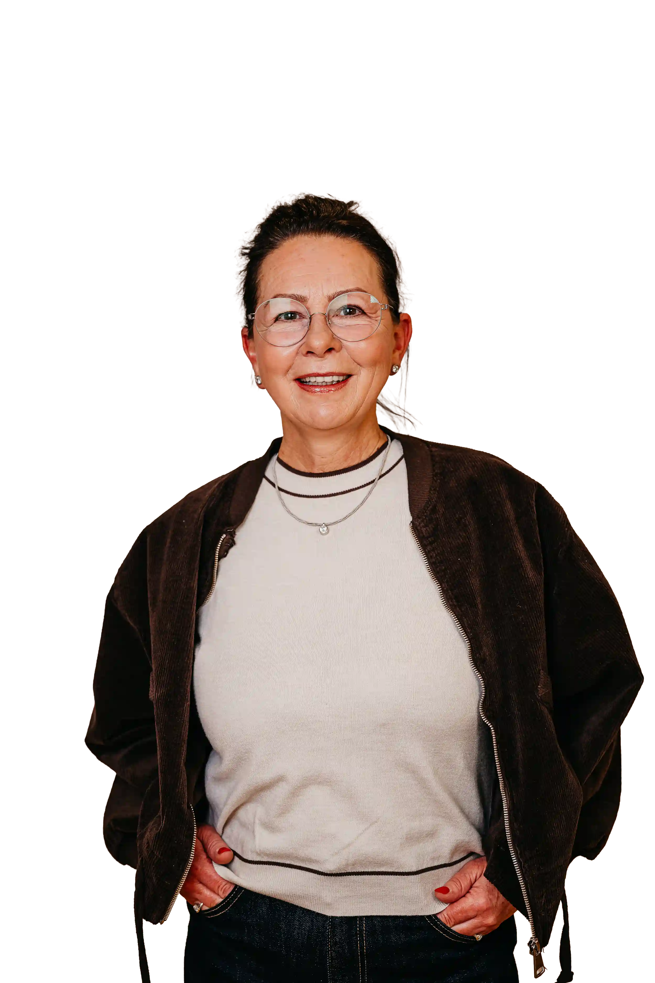 Fröhliche ältere Frau mit Brille, hellbeigem Pullover, brauner Jacke und dunkler Jeans, die die Hände in den Taschen hat.