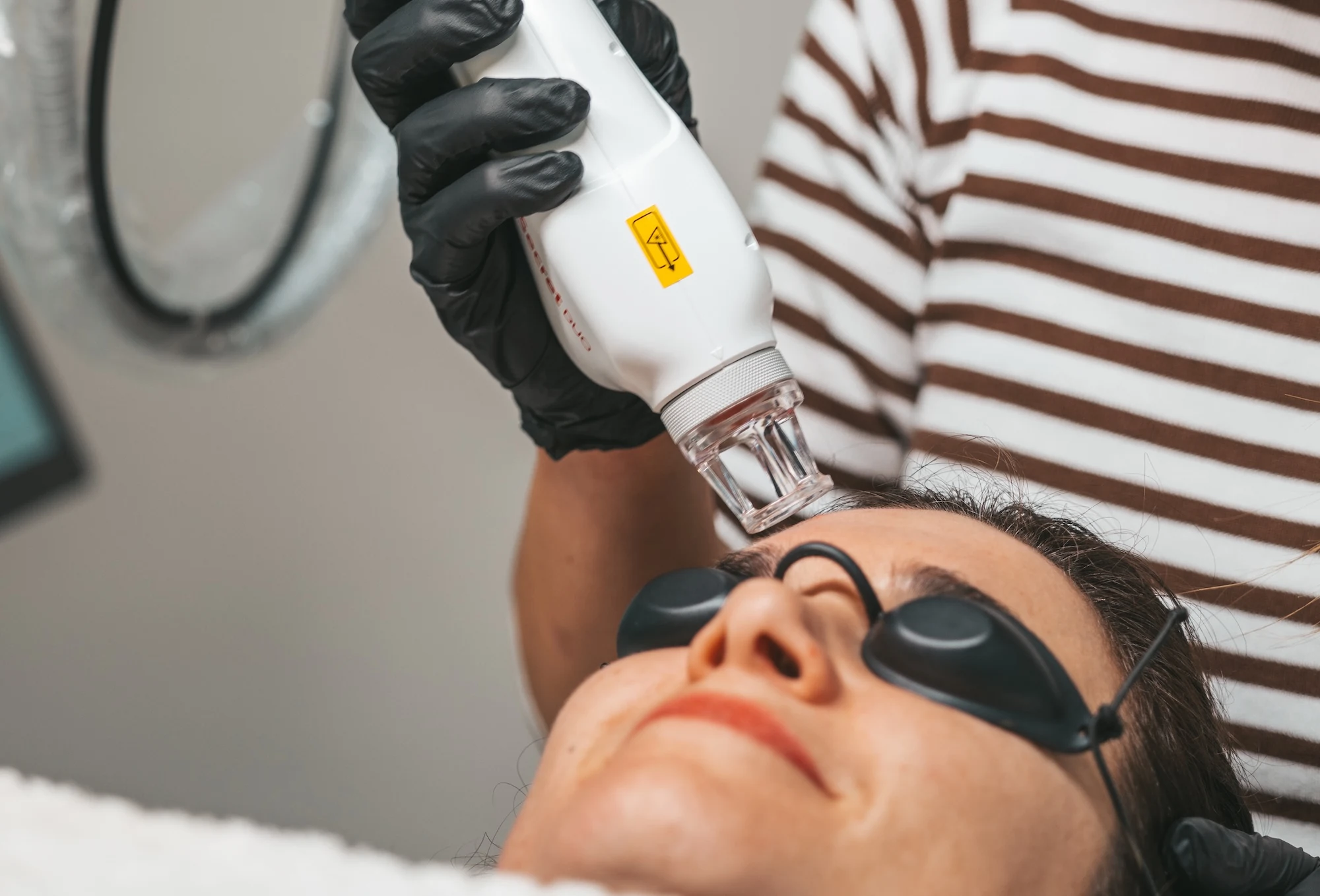 Frau trägt Schutzbrille, während ein Profi eine Hautbehandlung mit einem weißen Gerät am Gesicht durchführt.