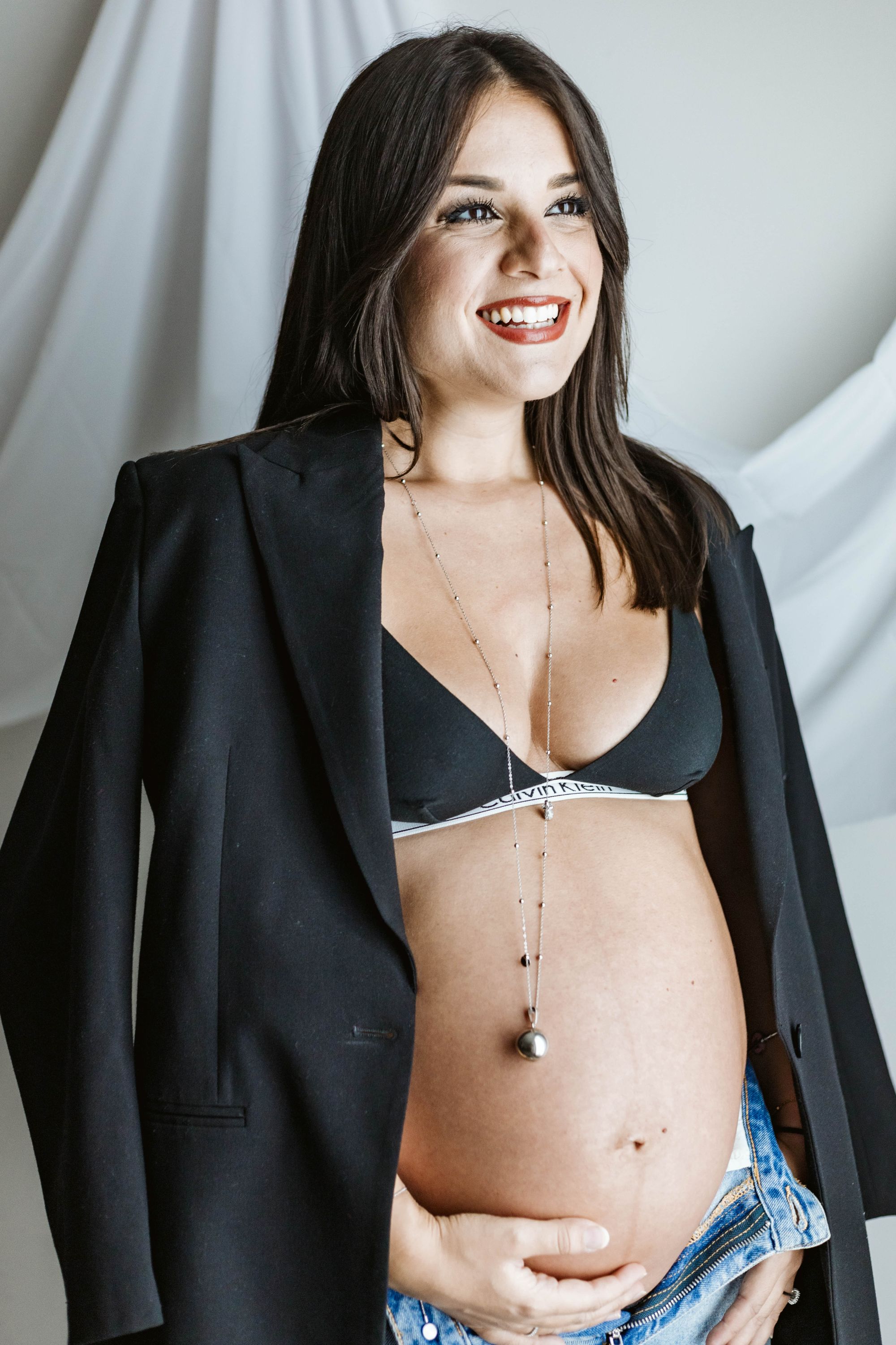 Smiling pregnant woman wearing a black bra, unbuttoned jeans, and a black blazer draped over her shoulders, with one hand on her belly.
