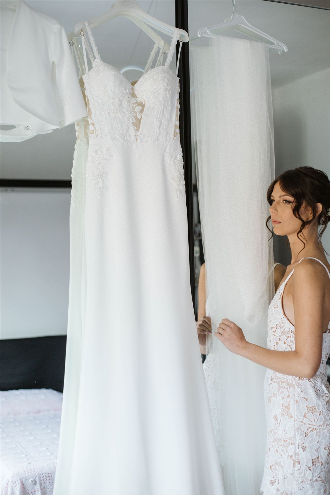 Woman in a lace slip dress stands beside a white wedding dress and veil hanging on a mirror.