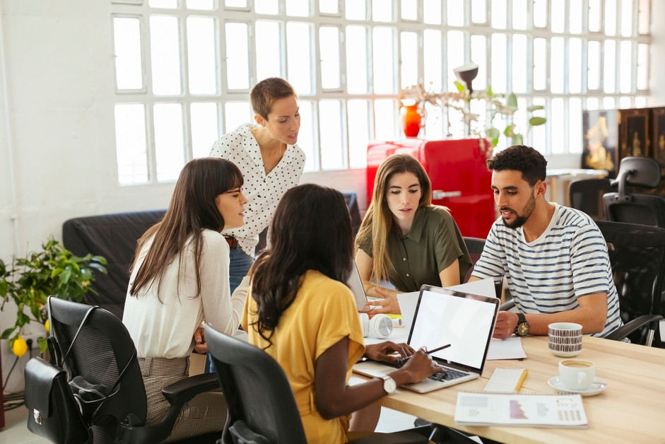 A group of people gather around a laptop at an office meeting table while looking for files.