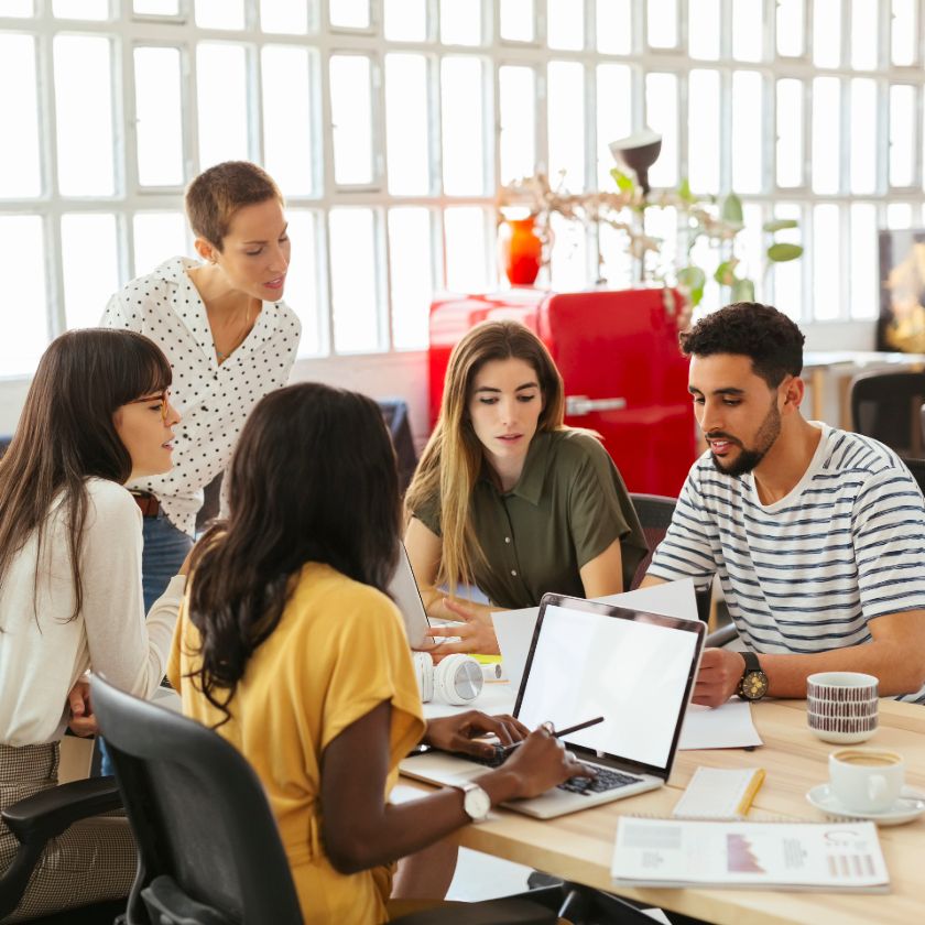 A group of people gather around a laptop at an office meeting table while looking for files.