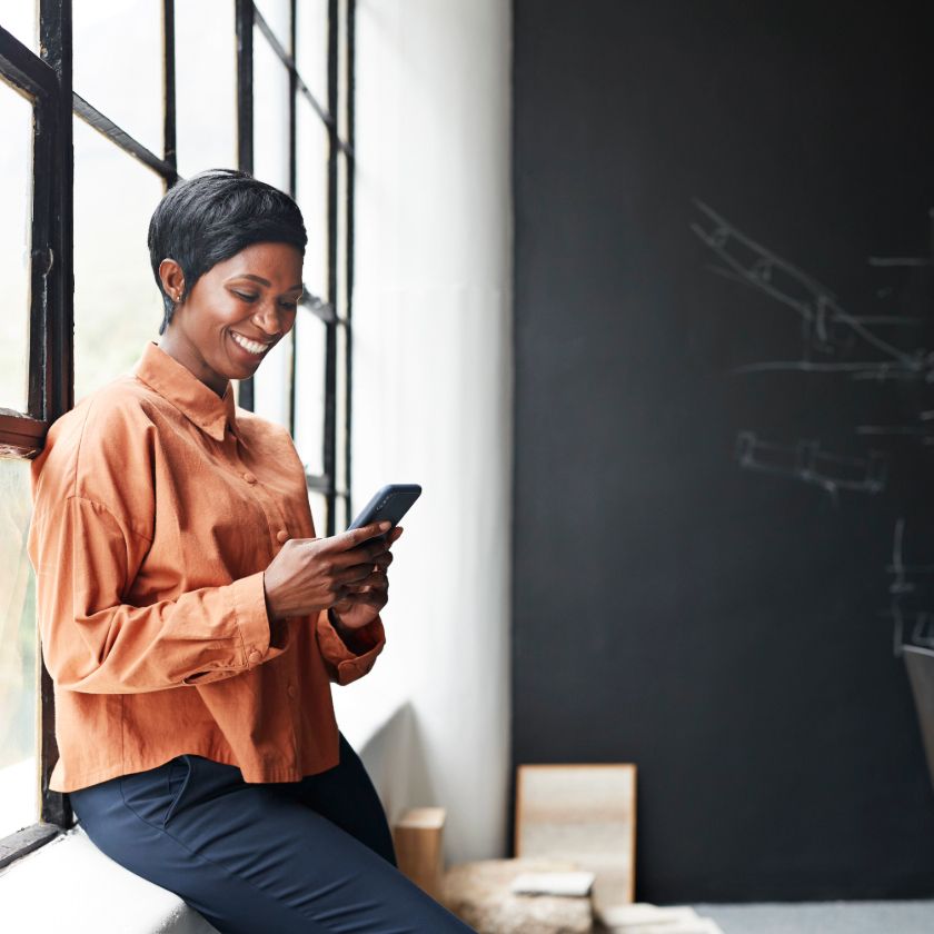 A person sits by a window in front of a blackboard while looking at their phone and smiling.