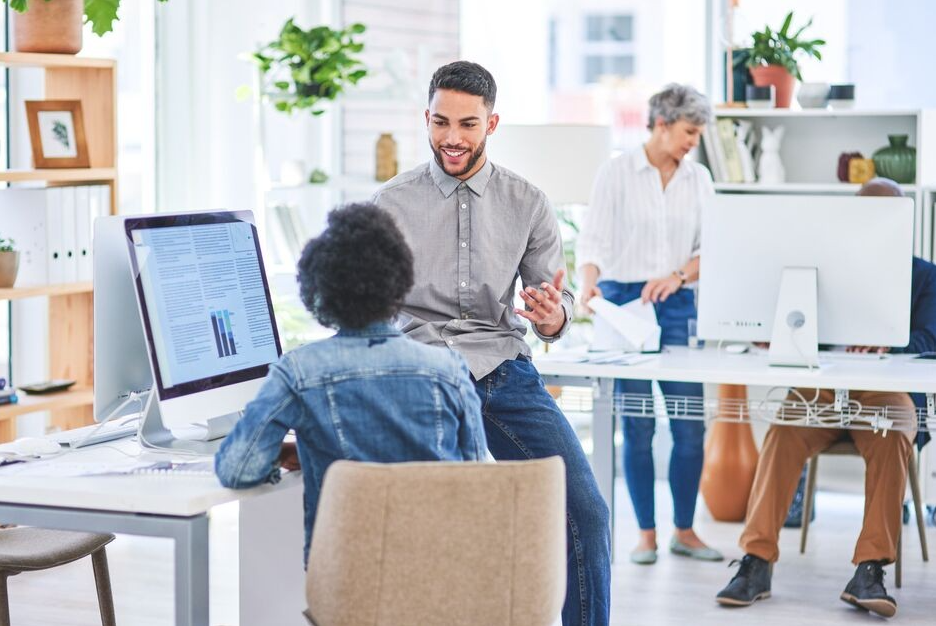 A person sits at a colleagues desk and has an enthusiastic work discussion.