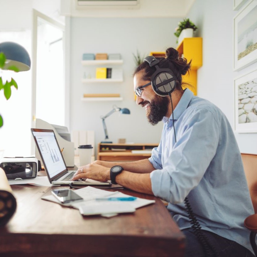 A person wearing headphones smiles while working on a laptop and looking at sales charts at a home desk, surrounded by documents.