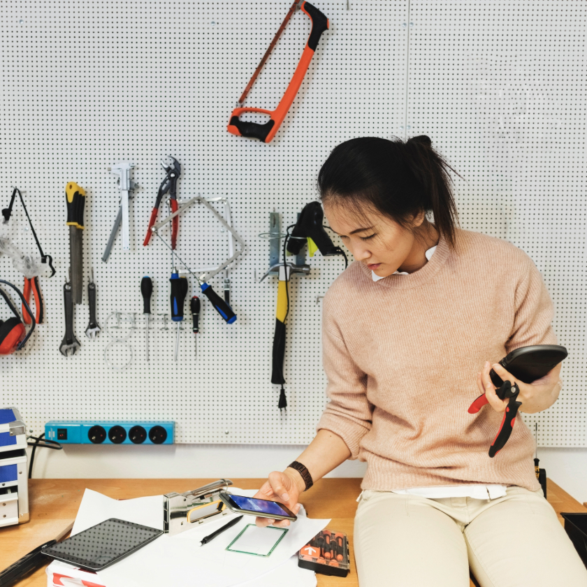 A person sits in a workshop surrounded by tools while checking their phone.