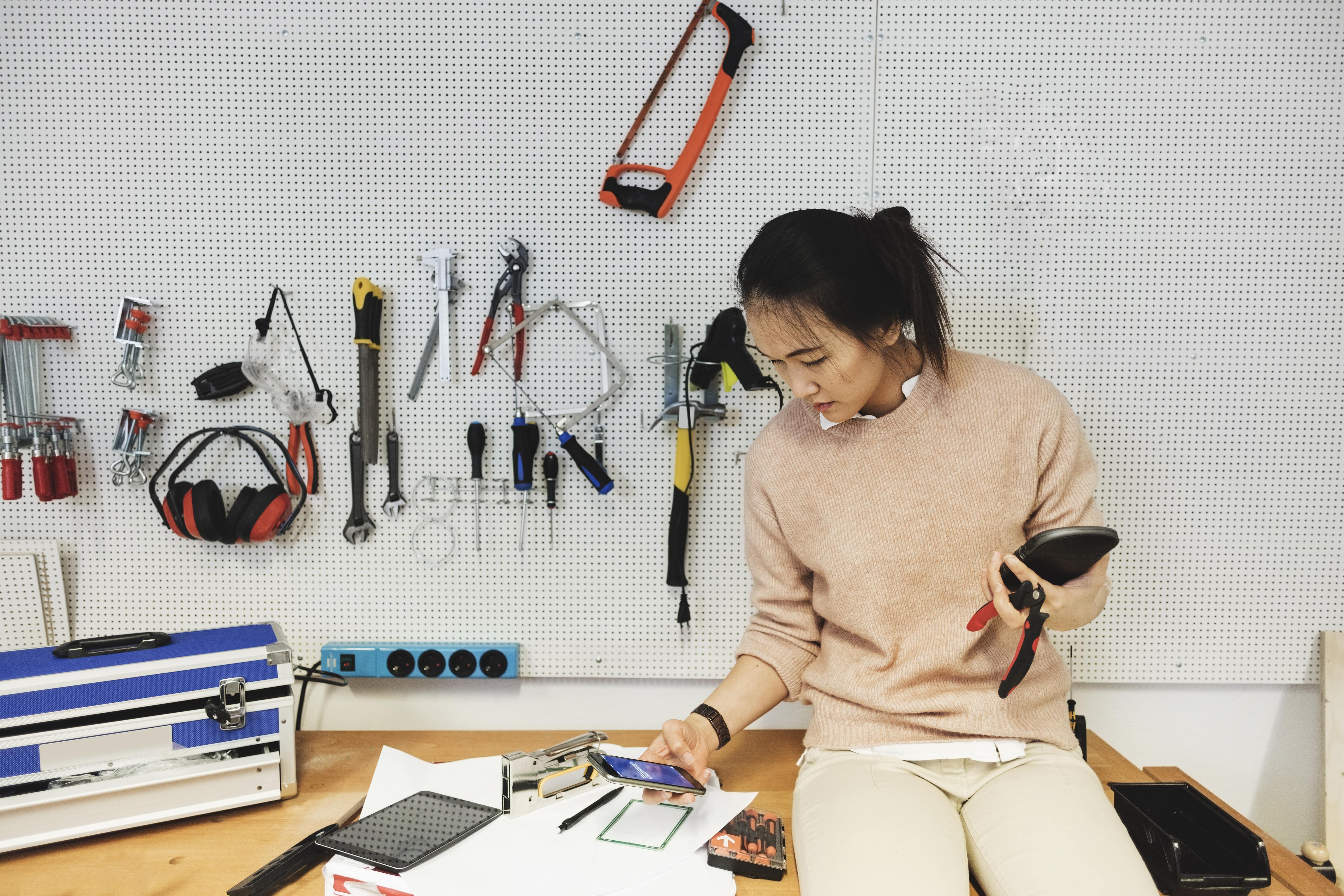 A person sits in a workshop surrounded by tools while checking their phone.