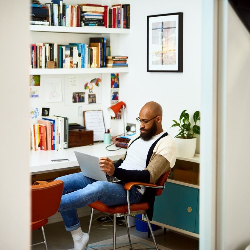 A person sitting at a desk in a home office, reviewing content on a laptop, surrounded by books and notes.