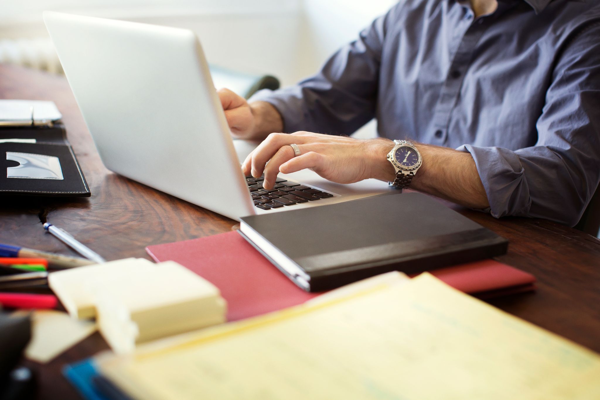 A person sits at their home desk while typing on a laptop surrounded by pens and papers.