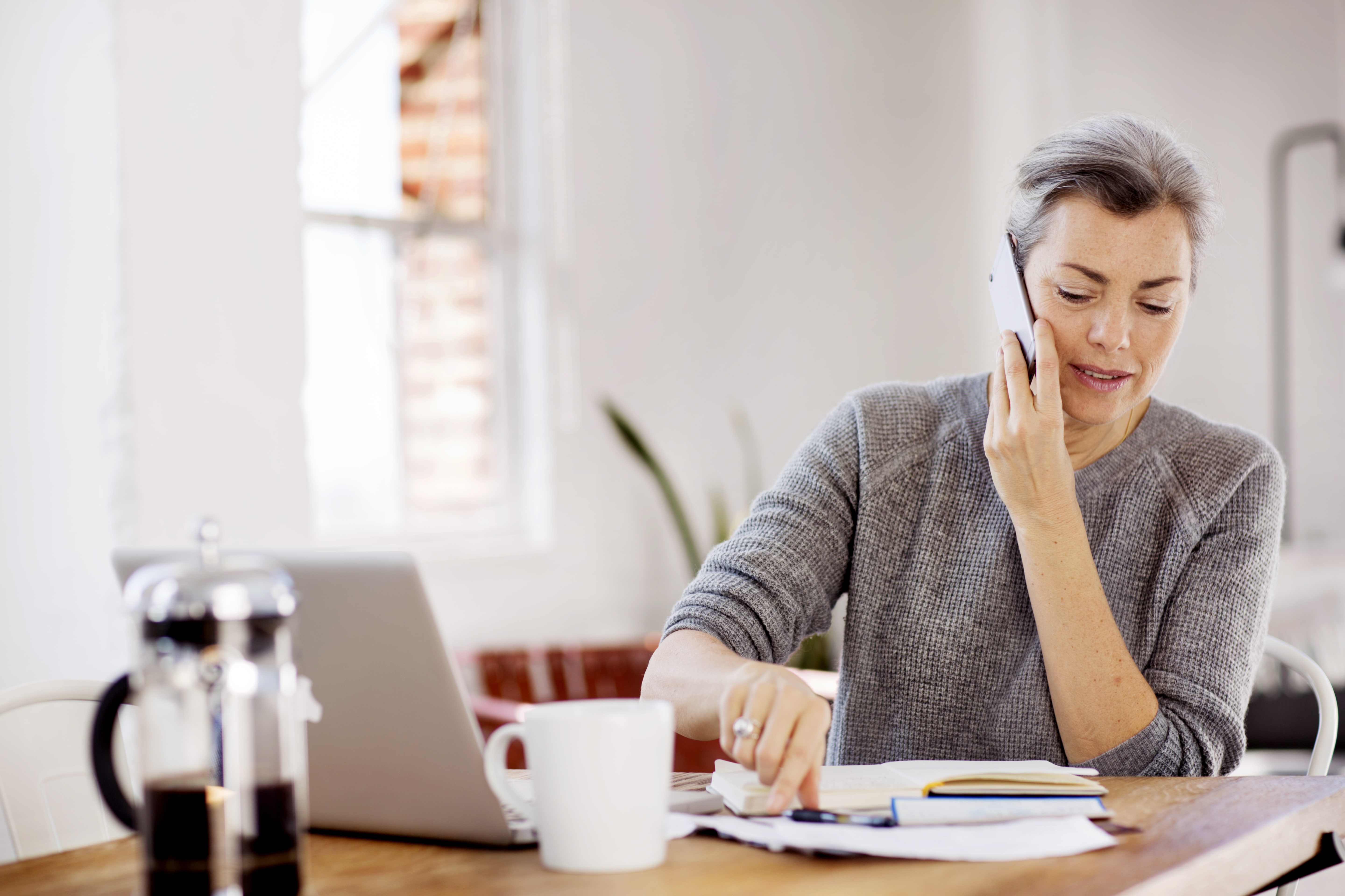 Marketing manager sits at desk on phone while reviewing notes next to laptop and coffee mug.