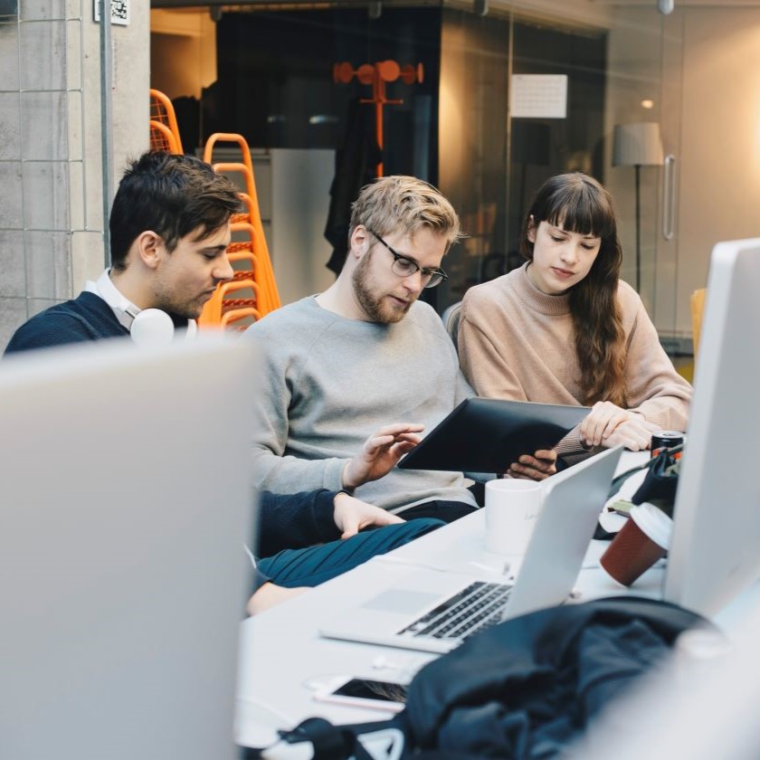 A group of colleagues gather in an open-plan office and discuss something while looking at a tablet device.