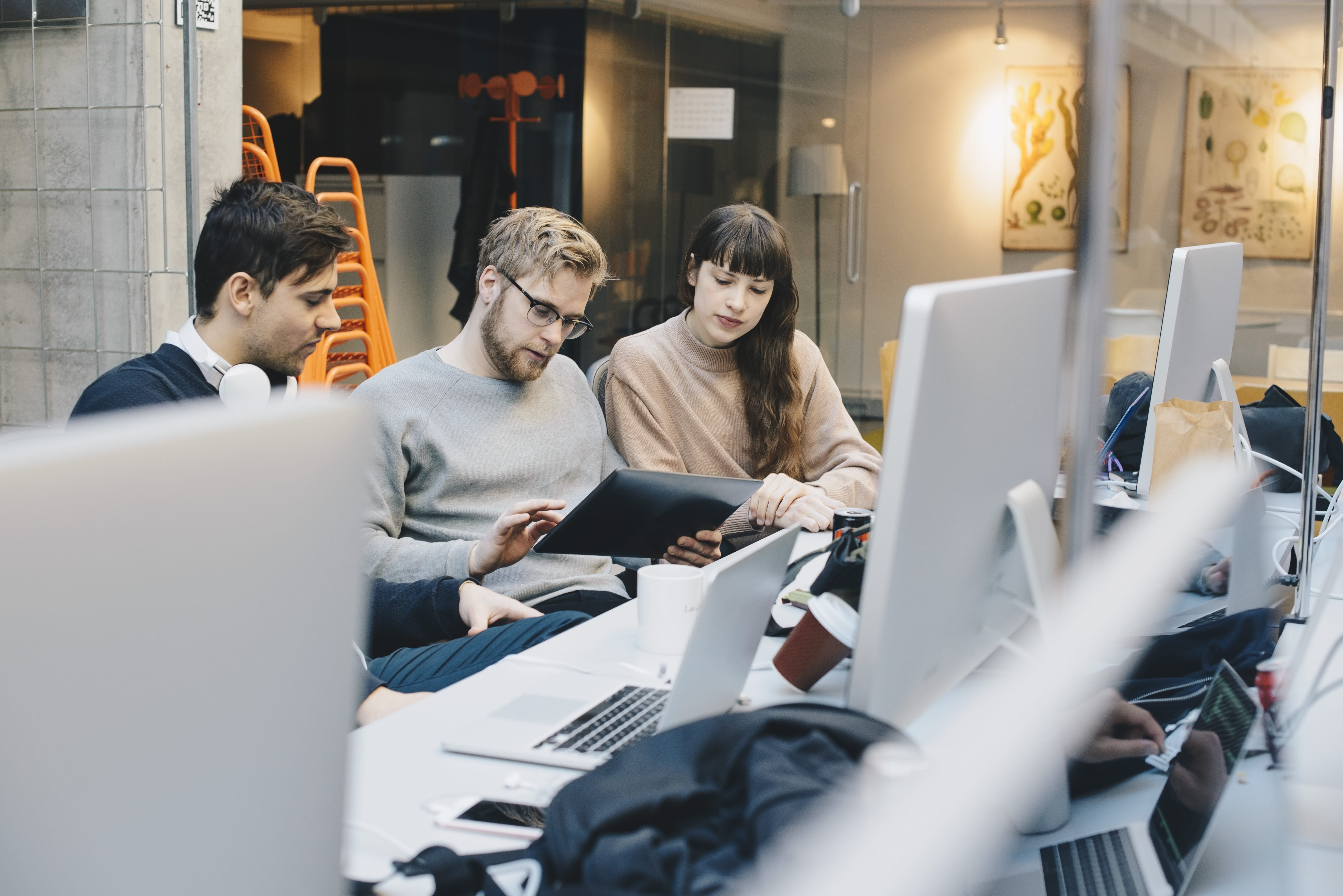 A group of colleagues gather in an open-plan office and discuss something while looking at a tablet device.