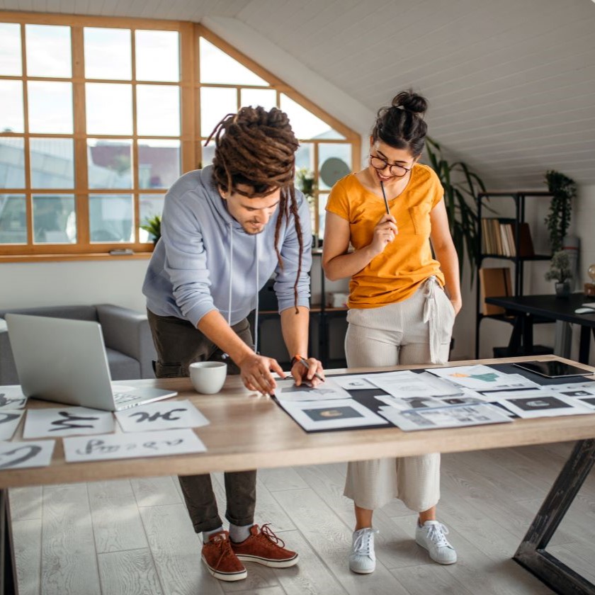 Designers evaluating printed marketing concepts on paper and laptop in a home studio with natural light and plants.