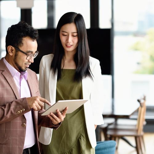 Two people stand in an office while consulting a tablet device.