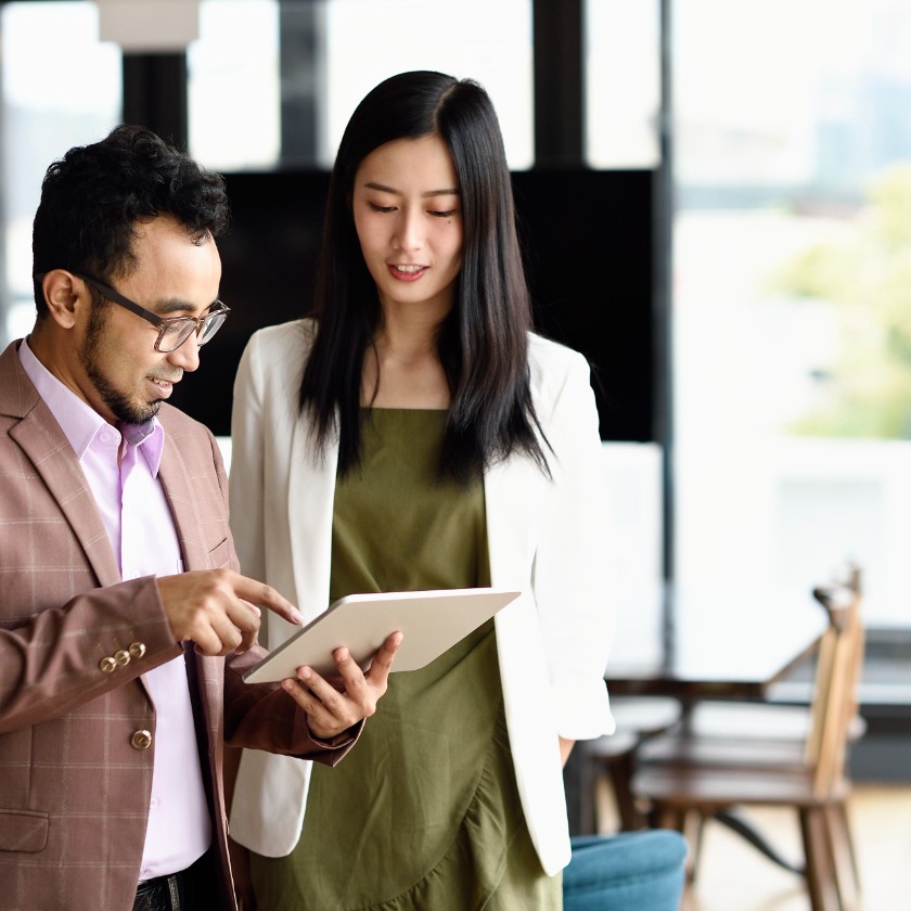 Two people stand in an office while consulting a tablet device.