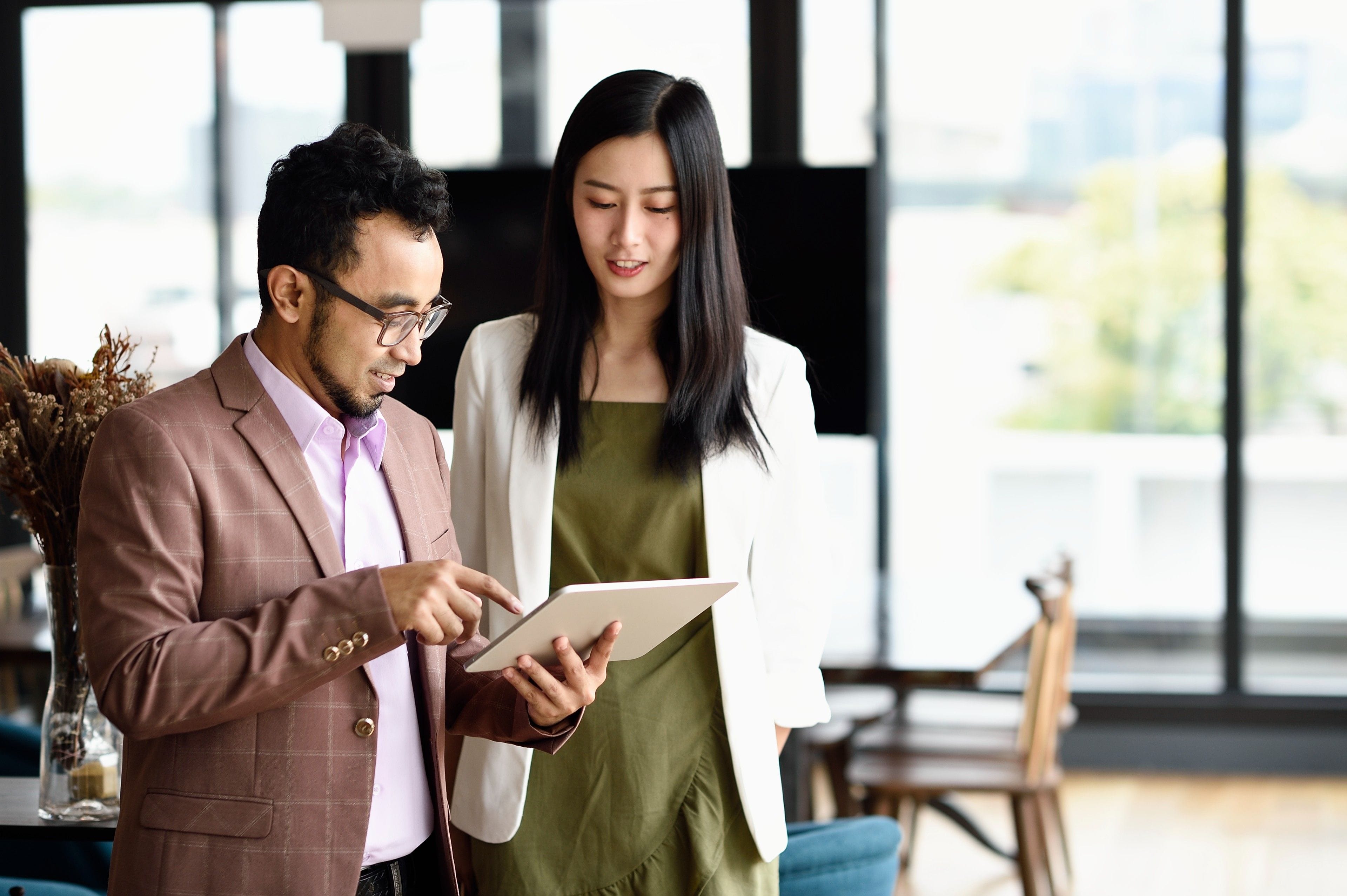 Two people stand in an office while consulting a tablet device.