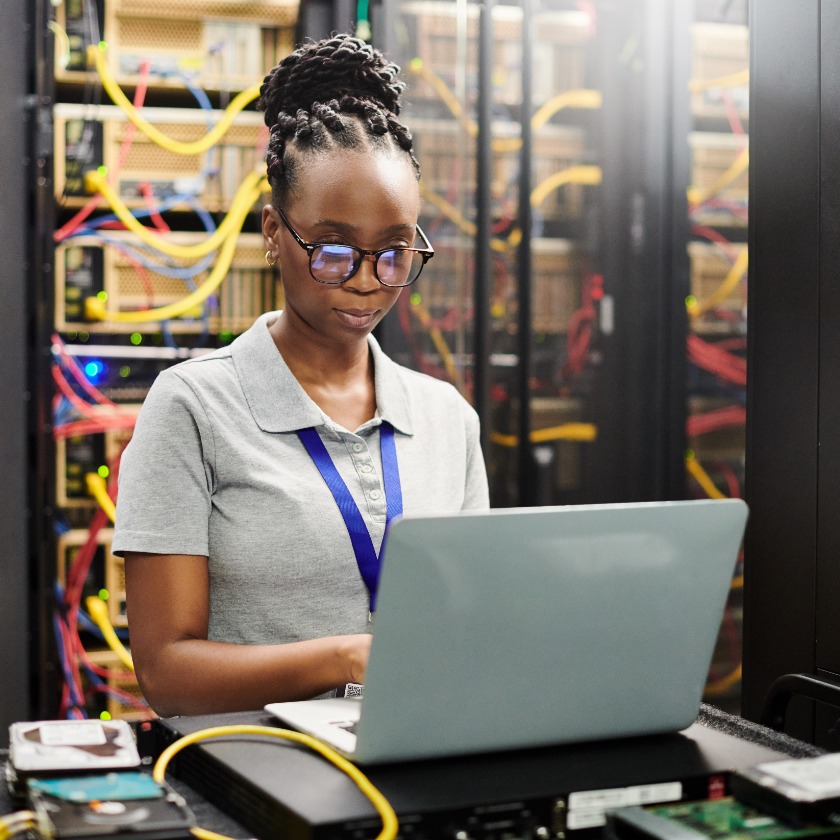 An engineer attends to a task on while working on a laptop in the middle of a large data center.