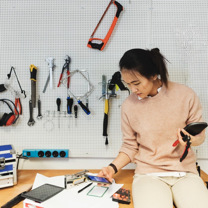 A person sits in a workshop while checking their phone and consulting some project plans.