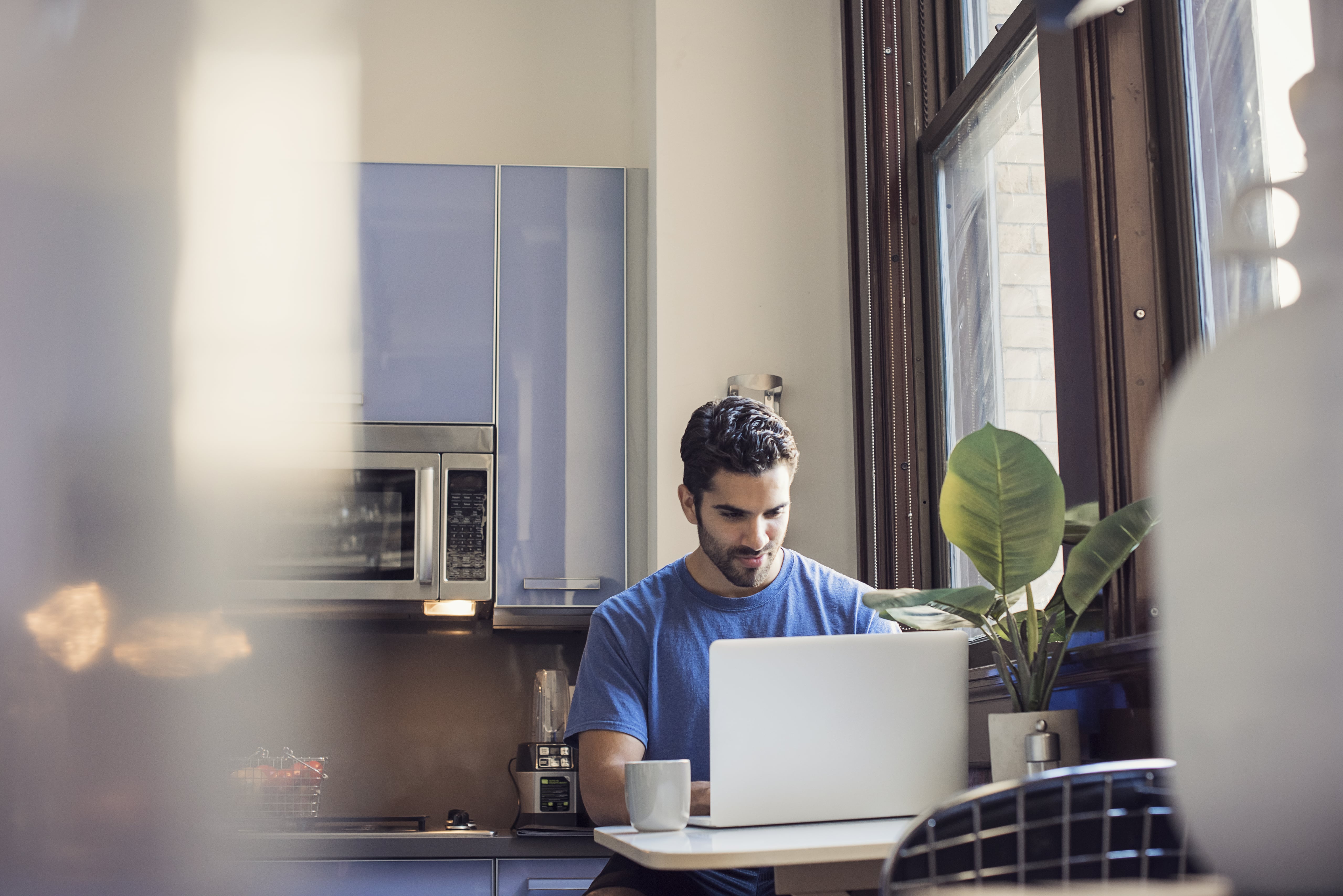 A person using a laptop near a window with natural light, plant and coffee cup on the table.