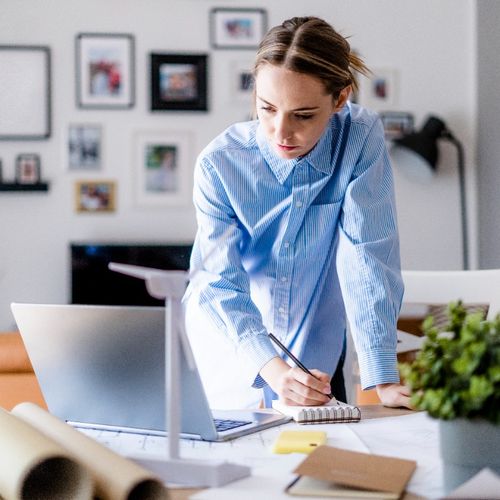 A person leans over their home office desk while taking notes on a pad and looking at their laptop screen.