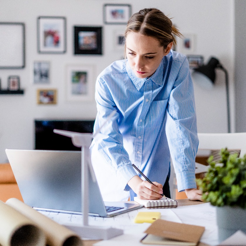 A person leans over their home office desk while taking notes on a pad and looking at their laptop screen.