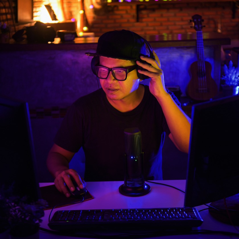 Person wearing headphones and a cap at a dual‑monitor computer, hand on mouse, with a desk microphone, under blue‑purple neon lighting.