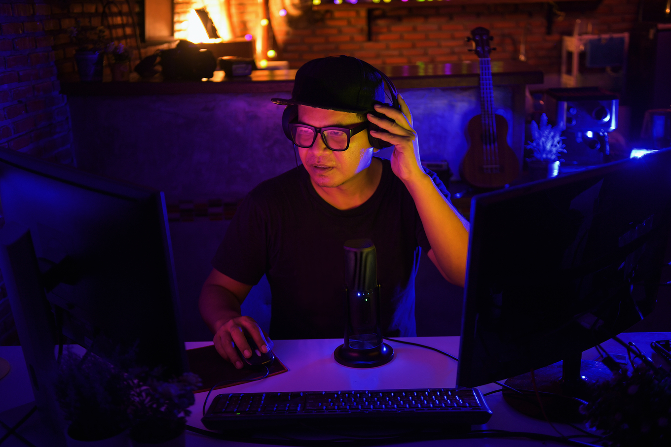 Person wearing headphones and a cap at a dual‑monitor computer, hand on mouse, with a desk microphone, under blue‑purple neon lighting.