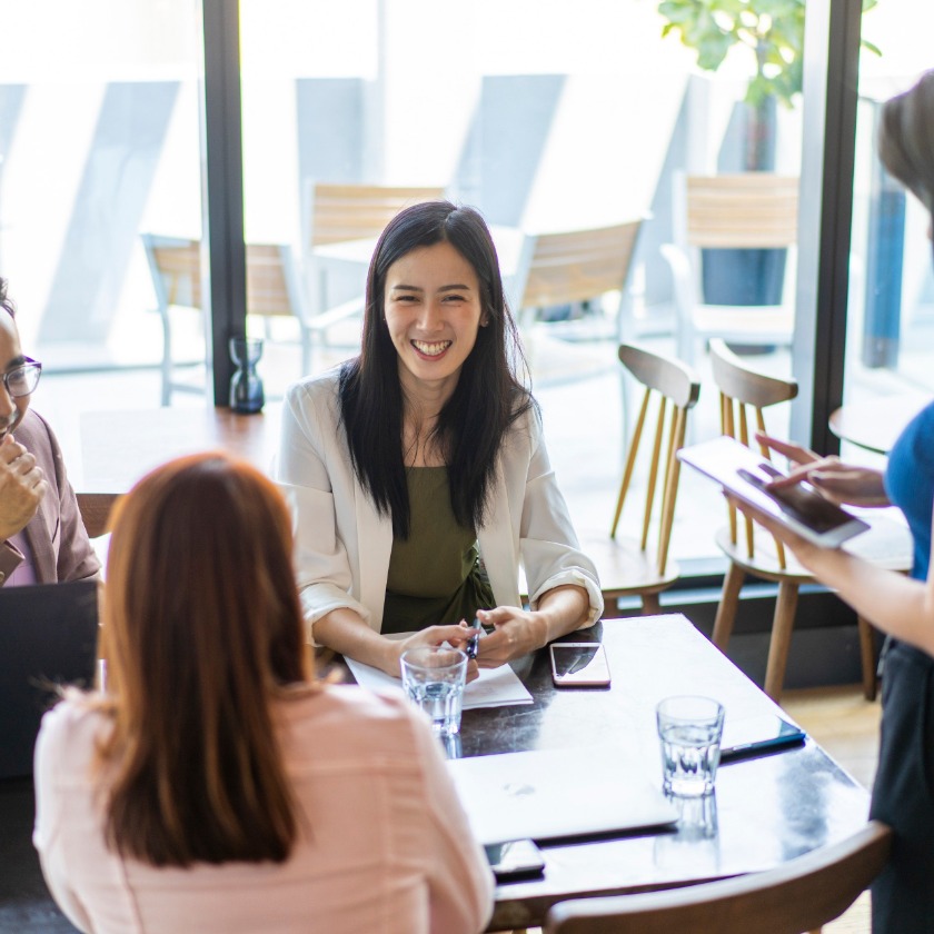 Smiling woman in a light blazer meets with colleagues at a café table by large windows, with laptops and water glasses.