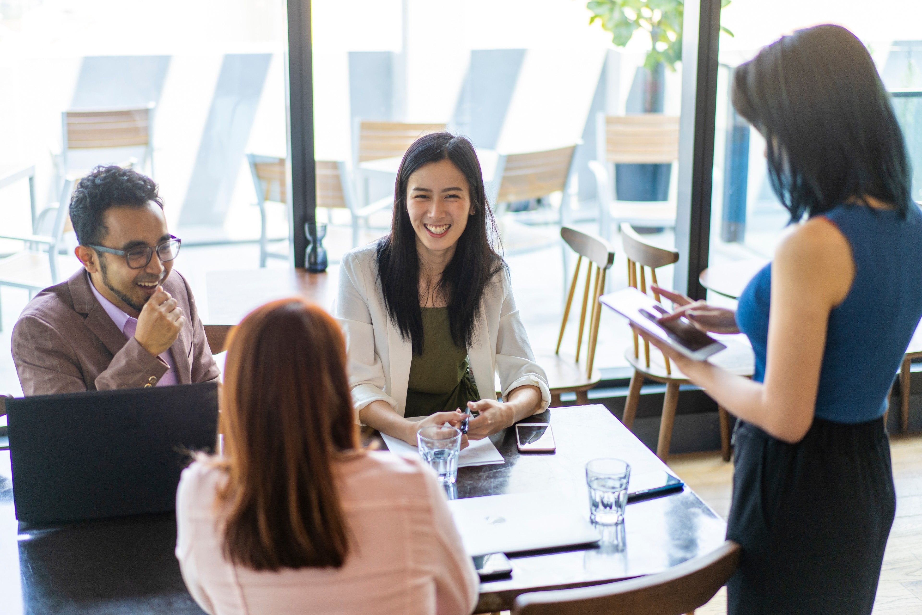 Smiling woman in a light blazer meets with colleagues at a café table by large windows, with laptops and water glasses.