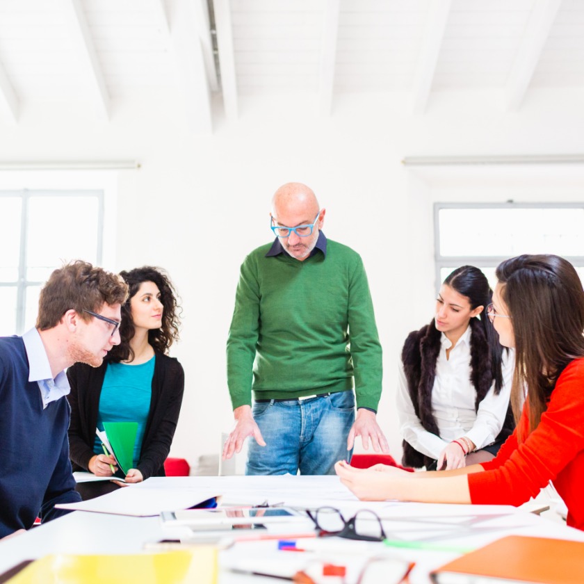 A team gathers around a meeting table covered in files while discussing a project.