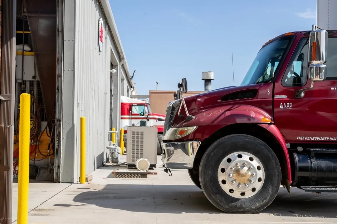 Technician measures semi-truck brake pad thickness with a caliper in an Omaha repair bay, with the exposed rotor, caliper, air lines, and suspension components visible.