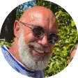 Foto de perfil circular de um homem sorridente com barba branca e cabeça raspada, usando óculos de sol escuros. Ele está ao ar livre, com árvores e folhagens verdes ao fundo.