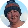 Foto de perfil circular de um homem sorridente usando um gorro de lã com listras nas cores cinza e laranja. O fundo é azul claro, sugerindo um céu ou uma paisagem ao ar livre.
