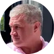 Foto de perfil circular de um homem de cabelos grisalhos curtos, olhando para o lado. Ele está vestindo uma camisa rosa clara e a imagem parece ter sido tirada ao ar livre, com um prédio e uma árvore desfocada ao fundo.