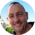 Foto de perfil circular de um homem sorridente com cabelos curtos e escuros, ligeiramente calvo. Ele está ao ar livre, com o céu azul e uma construção ao fundo, sugerindo um local turístico ou urbano.
