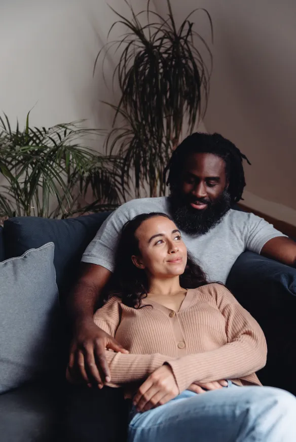 Imagem de um casal sentado em um sofá. A mulher, de cabelos longos e escuros, está encostada no peito do homem, que tem barba e cabelo crespo. Ambos estão em um ambiente aconchegante, com almofadas e plantas ao fundo, e aparentam estar relaxados e confortáveis.