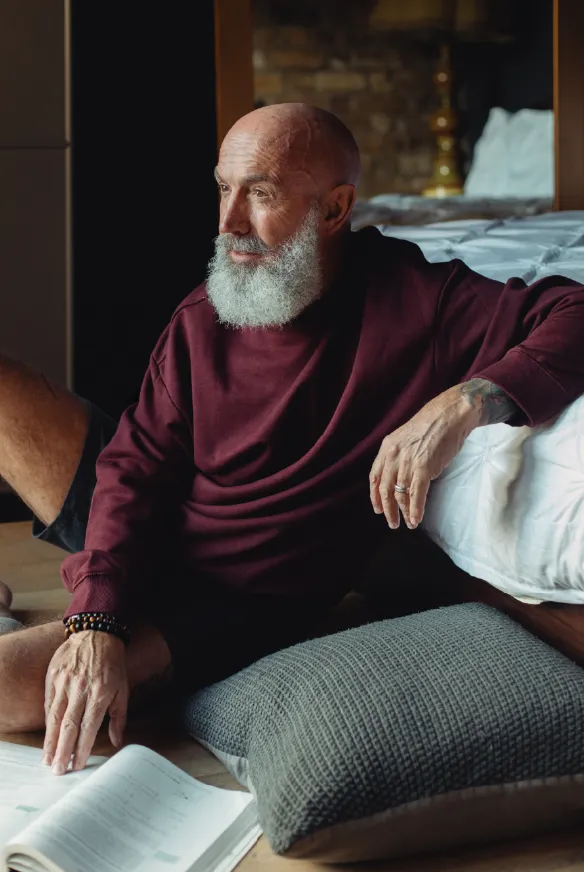 Imagem de um homem idoso com barba branca e cabeça raspada, vestindo um moletom escuro, sentado ao lado de uma cama em um ambiente com luz suave e decoração aconchegante.