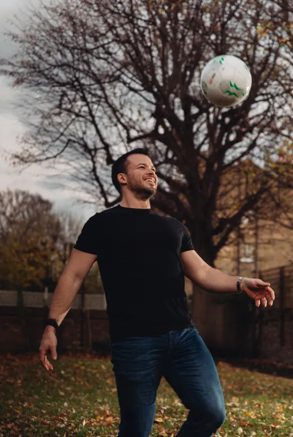 Imagem de um homem jovem vestindo uma camiseta preta e jeans, brincando com uma bola de futebol em um parque durante o outono. As árvores ao fundo têm folhas caídas, e a atmosfera é tranquila e descontraída.